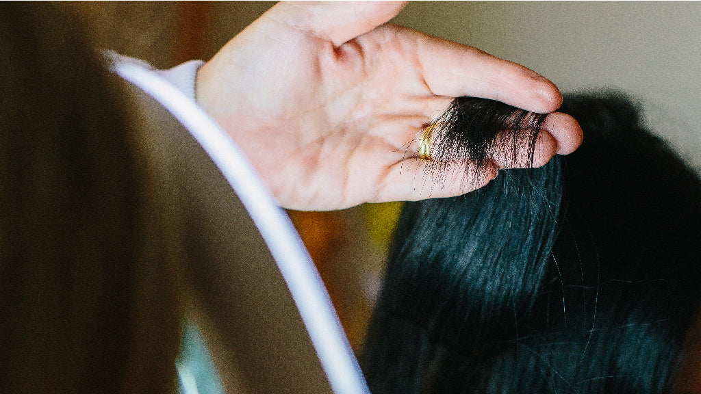 Close up photo of a white person's hand holding up some strands of long black hair while the person with the long hair sits in front of them, facing away from the camera