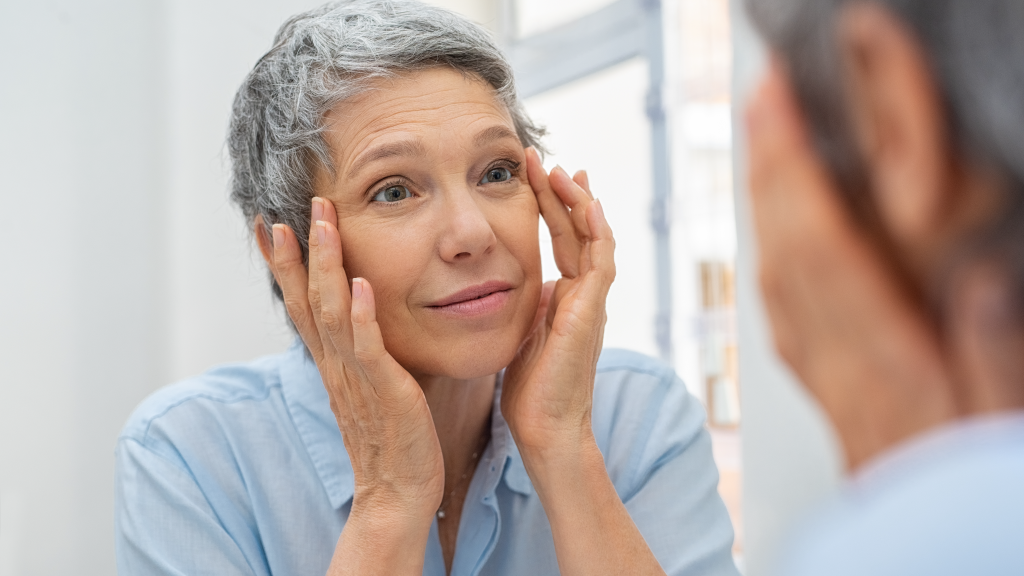 Photo of a white woman with short grey hair looking at her skin in the mirror